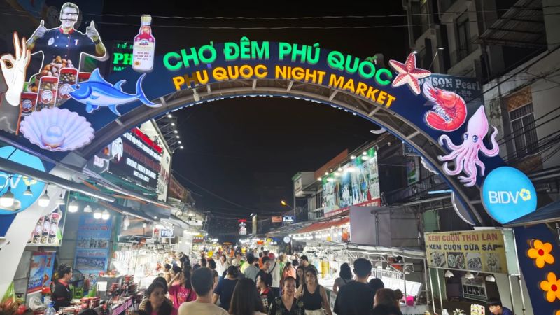 Cổng chợ đêm, phía trong nhiều gian hàng tập trung đông khách tham quan, mua sắm.